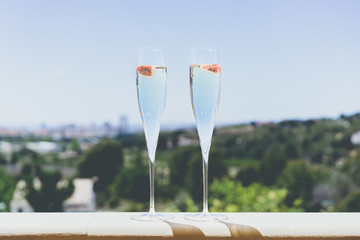 Two champagne glasses with strawberry on sunny terrace outdoor patio overlooking green trees at summer day outside of the city