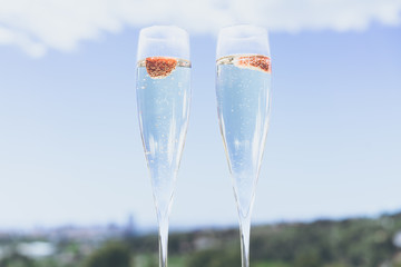 Two champagne glasses with strawberry on sunny terrace outdoor patio overlooking clean blue sky at summer day outside of the city