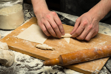 Chef hands are rolling a croissant from dough on wooden board. black background. close-up view