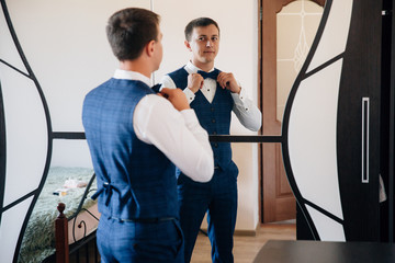 The guy dressed his business suit and tied a stylish bow-tie. The man gathers in front of the mirror and smiles at his reflection.