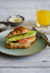 freshly baked croissant filled with salmon and green leaves and yellow cheese, placed on a mint green plate, served with orange juice and cream cheese on the side. all placed on a wooden white table.