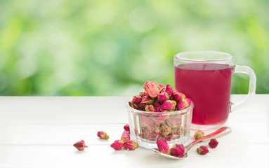Tea rose buds.Dry rose flowers in glass, tea in cup and spoon on white wooden board.Green bokeh on background