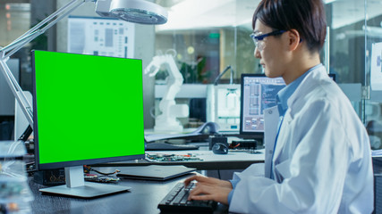 Asian Scientist Sitting at His Desk Works on a Personal Computer with Mock-up Green Screen. In the Background Computer Science Research Laboratory with Robotic Arm Model.