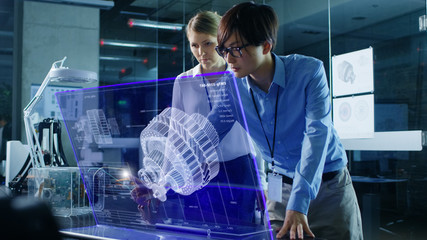 Male and Female Engineers Have Discussion while Using Modern Computer With Transparent Holographic Display. Monitor Shows Mechanical Gear Detail Visualization. Shot in Modern Glass Office.