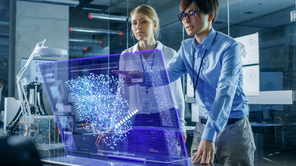 Male and Female Engineers Have Discussion while Using Modern Computer With Transparent Holographic Display. Monitor Shows User Neural Network Architecture Visualization. Shot in Modern Office.