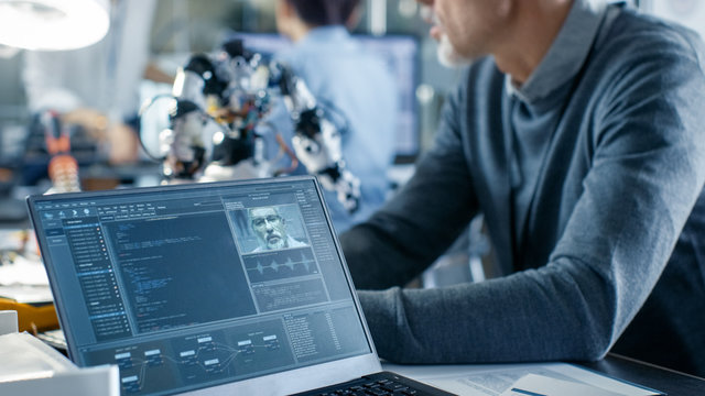 Robotics Engineer Manipulates Voice Controlled Robot, Laptop Screen Shows Speech and Face Recognition Software. In the Background Robotics Reseatch Center Laboratory.