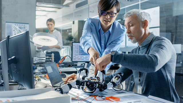 Two Engineers Talking while Working with Robot, Manipulating its Responses and Checking Data with Laptop Computer. In the Background Robotics Research Center Laboratory with Specialists Working.