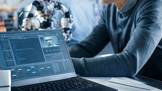Robotics Engineer Manipulates Voice Controlled Robot, Laptop Screen Shows Speech and Face Recognition Software. In the Background Robotics Reseatch Center Laboratory.