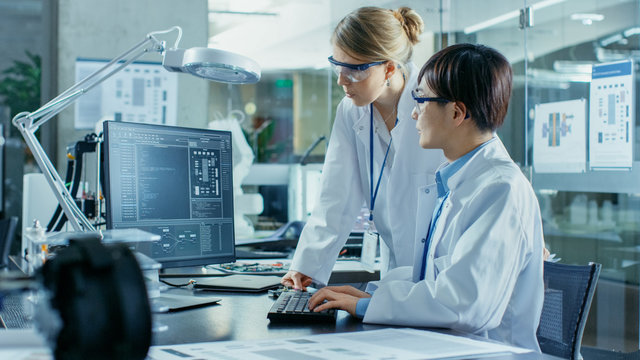Asian Scientist Sitting at His Desk Consults Senior Engineer about Sophisticated Coding and Programming. In the Background Computer Science Research Laboratory with Robotic Arm Model. - Powered by Adobe