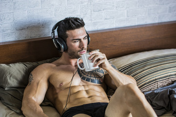 Shirtless muscular sexy male model listening to music on headphones and drinking tea or coffee, lying alone on bed in his bedroom, relaxing with coffee or tea cup