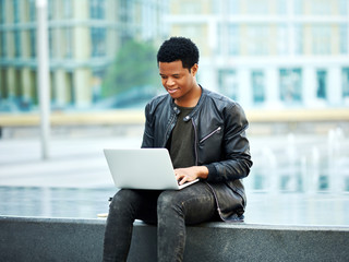 African American student sitting on stone seat and doing his homework on laptop computer outdoors in city