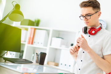 A young man in glasses stands near a computer desk. A young man draws a marker on a magnetic board. On the neck, the guy's headphones hang.