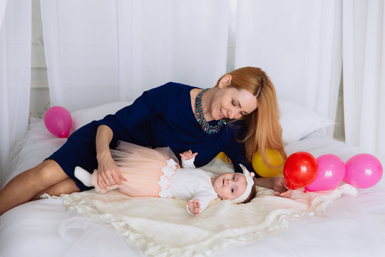 The child lies on the bed on his back and looks around, looking at the balloons on the bed. The girl's mother is lying on the side and gently hugs the baby.