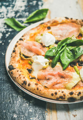 Italian lunch or dinner. Freshly baked pizza with artichokes, smoked turkey ham, olives, cream cheese and basil leaves over rustic wooden background, selective focus, close-up