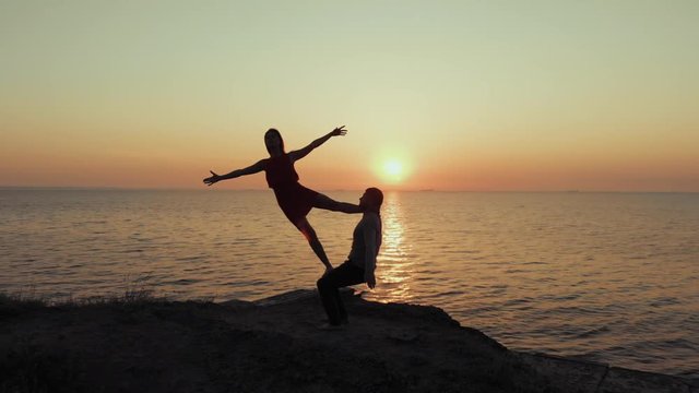 Silhouette of young woman and man doing pair yoga on sea beach at sunset. Meditation. Couple practicing acro yoga. Flexibility workout at nature background. Drone aerial view.