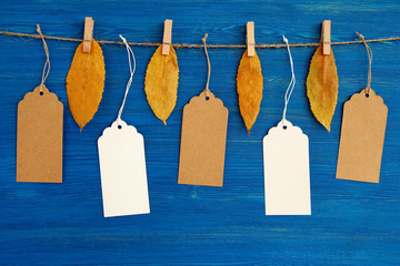 Five brown and white blank paper price tags or labels set hanging on a rope with dry yellow autumn leaves on the blue wooden background.