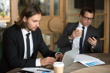 Young male businessman working at laptop, browsing internet, reading online news, colleague using smartphone surfing web or writing business email during company break in office. Technology concept
