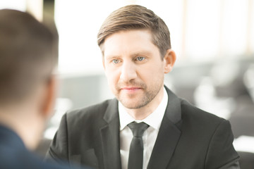 Young serious businessman looking at his colleague during conversation at meeting
