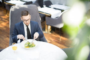 Contemporary businessman sitting by table in cafe or restaurant and eating vegetarian salad with juice