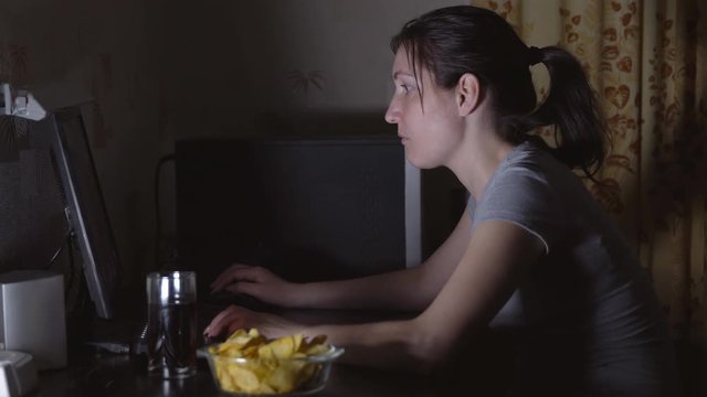 Young woman sitting at the computer and eating chips.