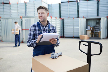 Young worker of transportation and storage service making notes in documents while standing by stack of large carton boxes