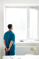 Rear view of young vet clinician in uniform standing by window and looking through it