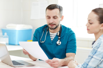 Obraz premium Young man in uniform of clinician showing medical document to his patient and consulting her about some details