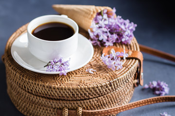 Fashionable rattan bag, cup of coffee and purple lilac in waffle cone on blue concrete background. Spring Summer Card Concept. Top view