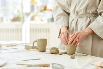 Obraz na płótnie Canvas Hands of female making mug from grey clay on table with supplies for creativity