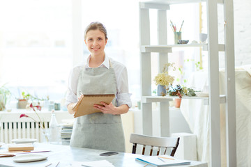 Happy craftswoman standing by workplace, making sketches in notepad and looking at camera
