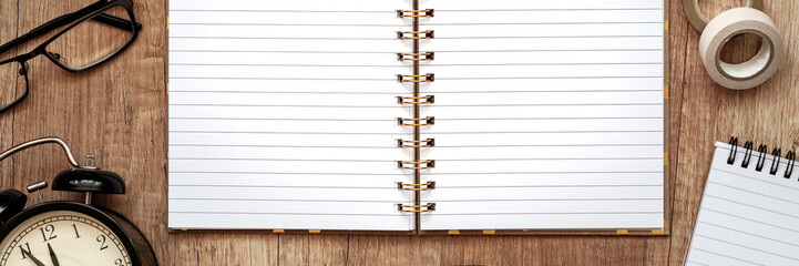 Panoramic view of a wooden desk from above with an open notebook, glasses and a clock