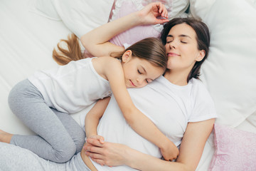 Obraz na płótnie Canvas Pleased little child sleeps near her mother, embraces with love, has pleasant dreams, lie on comfortable bed. Mum and cute daughter have good sleep in bedroom. Family, sleeping and rest concept