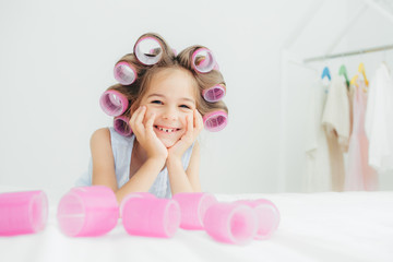 Obraz na płótnie Canvas Portrait of cheerful female kid keeps hands under chin, has curlers on hair, going to have nice hairstyle, poses against white background, has charming smile, being in good mood. Children and beauty