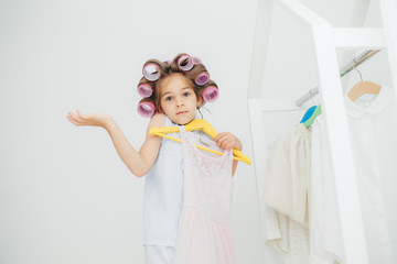 Hesitant girl with clueless expression, doesn`t know what to wear, holds hangers with dress, has curlers on head, isolated over white background. Cute child with pleasant appearance prepares for party