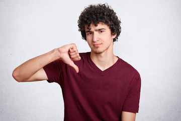 Portrait of attractive handsome male with curly hair, shows dislike gesture, keeps thumb down, wears casual t shirt, isolated over white background. Sullen European man being discontent by something