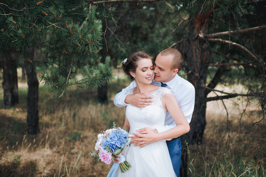 Beautiful and lovely newlyweds gently embrace in a green pine forest on a sunny day. A young groom hugs and kisses behind a beautiful bride in a lace dress in nature.