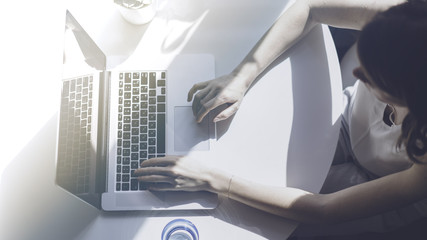 Young positive beautiful woman working on a laptop sitting at the table in living room of modern house.Top view. Flares
