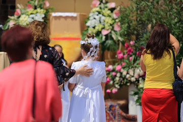 Dziewczynka w białej sukience komunijnej i wianku stoi przed ołtarzem z kobietami.