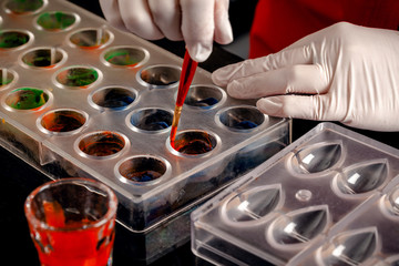A woman confectioner with red uniform and white sterile gloves do a set of colorful chocolates from milk chocolate on a table.