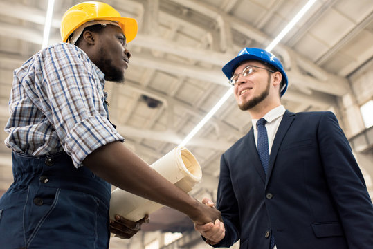 Low angle view of confident African American engineer wearing hardhat and overall shaking hand of his smiling superior after successful completion of promising project discussion