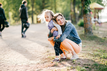 Naklejka na ściany i meble Happy smiling mother hugging her lovely little daughter outdoor. Lifestyle family. Adult cheerful female parent playing with her beautiful emotional child at nature in summer. Positive people faces.