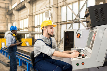 Bearded worker wearing protective helmet and overall operating machine while his African American colleague controlling manufacturing process, interior of production department on background