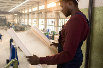 Profile view of handsome African American engineer wearing overall and protective helmet studying blueprint with concentration while standing at production department of modern plant