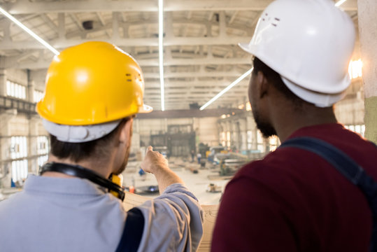 Rear view of bearded engineers wearing hardhats and overalls brainstorming on promising joint project while standing at spacious production department of modern plant with blueprint in hands.