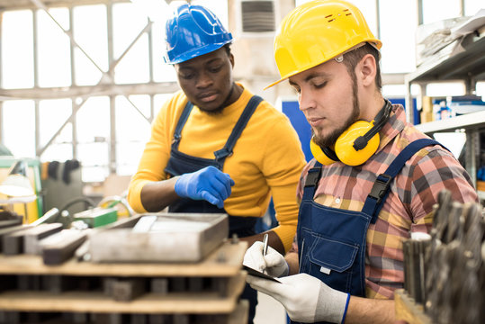 Bearded inspector wearing hardhat and overall taking notes while carrying out quality control at production department of modern plant, African American machine operator answering his questions