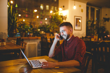 Young handsome Caucasian man with beard and toothy smile in red shirt works behind laptop, hands on keyboard sitting at wooden table. Uses calls on mobile phone. In evening at the coffee shop.