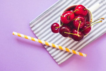 Close up and top view of fresh red cherries in a white cup on a gray and white stripped cloth, a yellow and white straw on pink or purple background. Hello summer, weight loss or healthy diet concept.