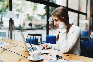 Fototapeta premium Hipster girl scrolling websites while surfing the internet online on a portable computer during morning coffee break. Business woman working on a laptop while having breakfast in a coffee shop.