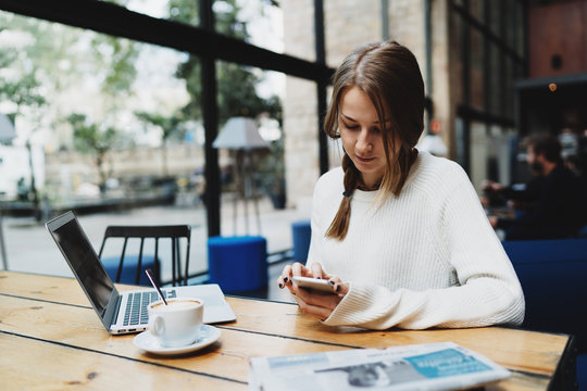 Half length portrait of a beautiful caucasian female with long blonde hair touching the screen of a mobile phone while sitting at the table with open laptop and morning newspaper on it.