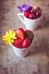 Close up and top view of fresh red cherries and strawberries in white cups decorated with yellow and purple flowers on an rustic wooden background. Hello summer, healthy diet or weight loss concept.
